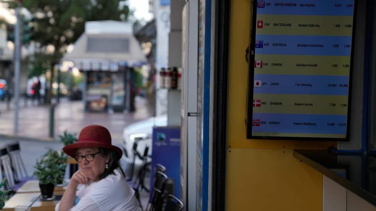 A woman sits at a cafe as a screen of a currency exchange office shows the foreign exchange at Omonia square in Athens, Greece, Wednesday, July 13, 2022. The euro on Tuesday fell to parity with the dollar for the first time in nearly 20 years. (AP Photo/Thanassis Stavrakis)